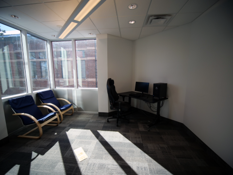 Bookable study room with corner windows; gaming PC with desk; two comfortable chairs; grey carpet. 